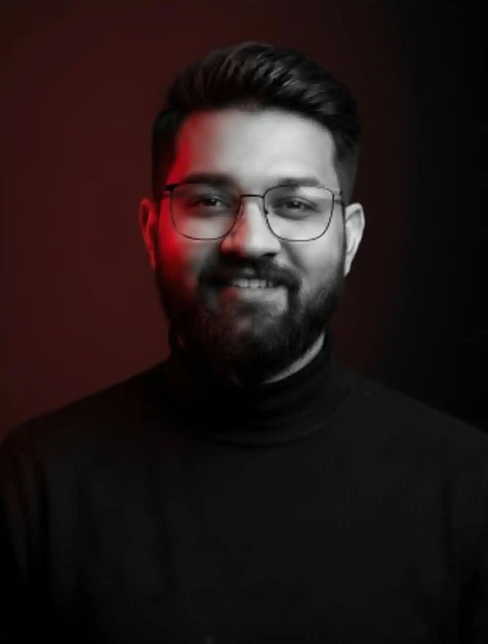 Portrait of a young man with glasses, smiling confidently against a dark background.